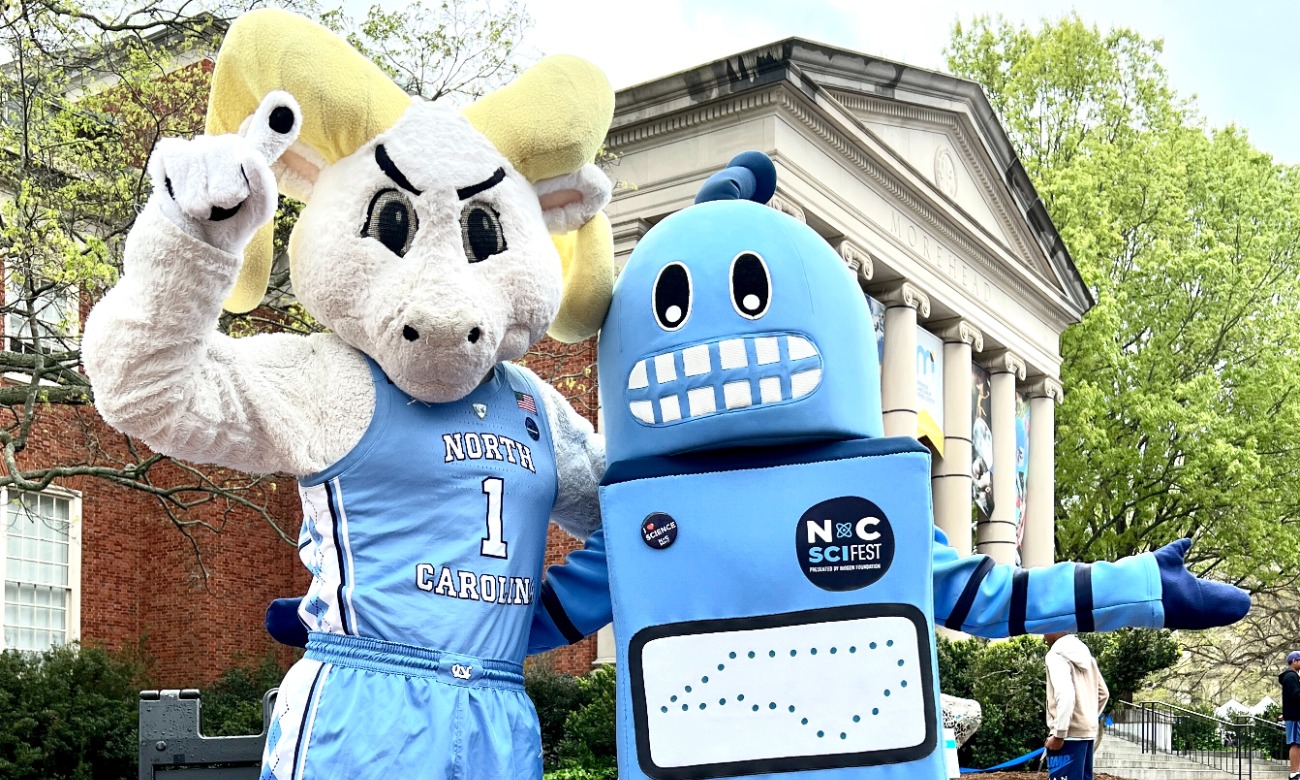 Ramses, the ram mascot, and a Kelvin, a blue robot mascot, are stood waving in front of the Morehead Planetarium,
