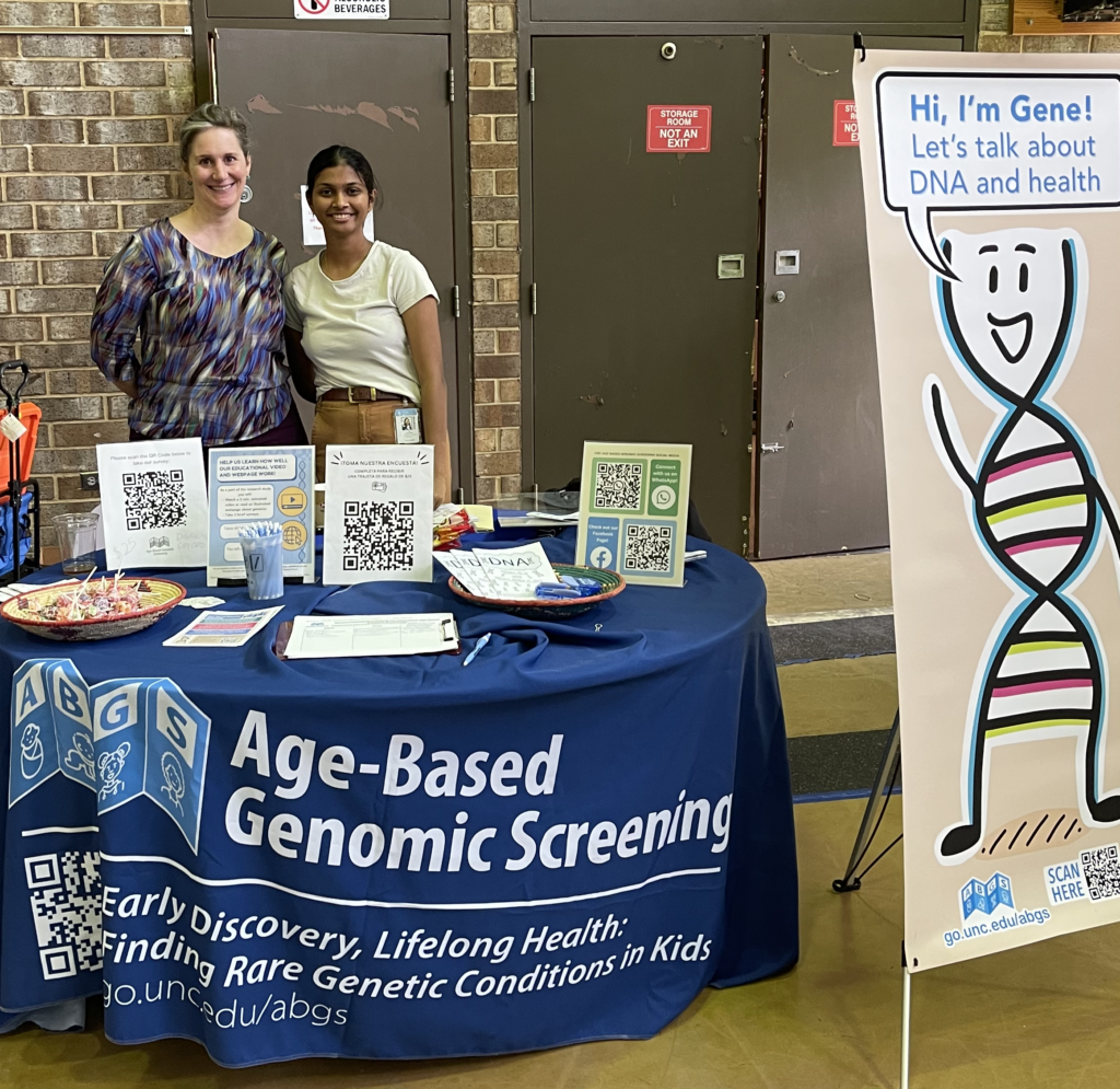 Laura Milko and Juhi Salunke at the Age-Based Genomic Screening Tabel with a standee of a DNA character beside them