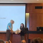 Selin Altinok receives her Pharmacology white coat from Dr. Henrik Dohlman on stage in front of her slide show.